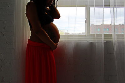 Midsection of pregnant woman standing by window at home