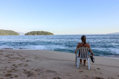 Rear view of woman sitting on chair at sandy beach against clear sky