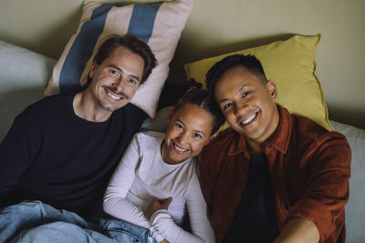 Directly above portrait of gay couple reclining with daughter on sofa at home