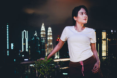 Young woman looking away while standing against illuminated buildings in city at night