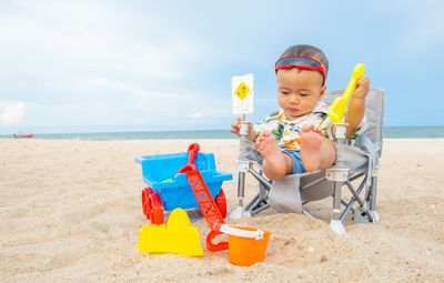 Boy sitting on chair at beach against sky
