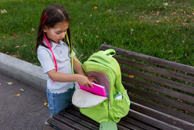 Young girl packing backpack on park bench for school day preparation