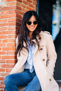 Portrait of smiling young woman wearing sunglasses against brick wall