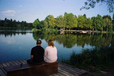 Rear view of couple sitting by lake against sky