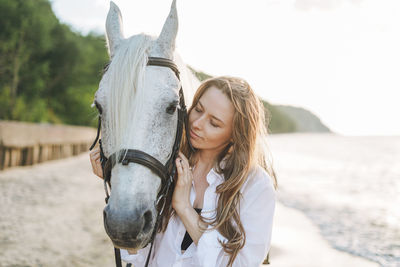 Young long hair woman in white shirt with white horse on seascape background