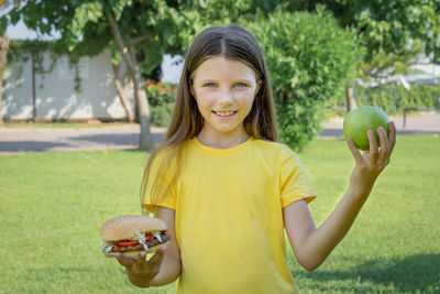 A teenage girl chooses between a burger and an apple outdoors in the park.