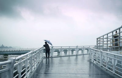 Man with umbrella standing on bridge against sky during rainy season