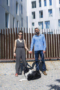 Full length portrait of couple with dog holding hands against building