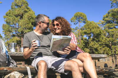 Smiling woman looking at man sitting with coffee and map on picnic table