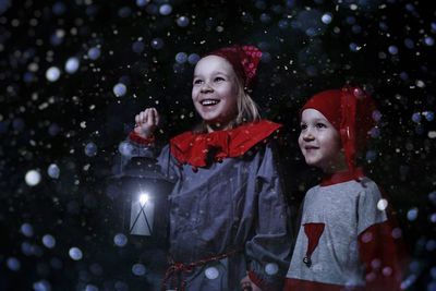 Cheerful siblings holding lantern while standing in snowfall during christmas