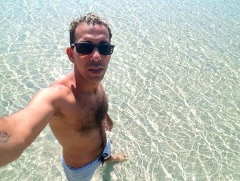 High angle portrait of shirtless man wearing sunglasses while standing in sea during sunny day