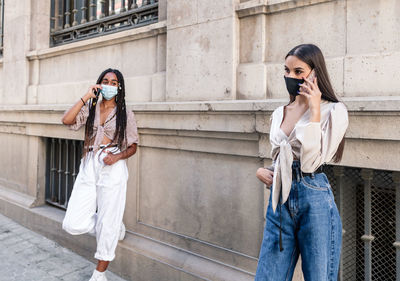 Serious multiracial females wearing trendy clothes and protective masks standing near building on street and speaking on cellphones during coronavirus pandemic