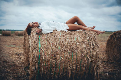 Side view of young woman sleeping on hay bale against cloudy sky at farm