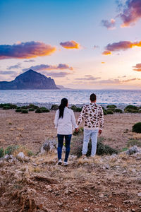 Rear view of men standing at beach against sky during sunset