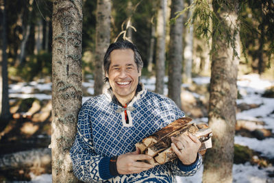 Portrait of happy mature man with firewood standing by tree in forest