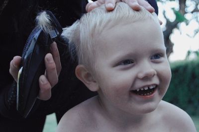 Midsection of person cutting boy hair smiling boy against trees