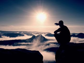 Silhouette of alone hiker in black and cap sit on cliff. blue filter photo. man looking to mist 