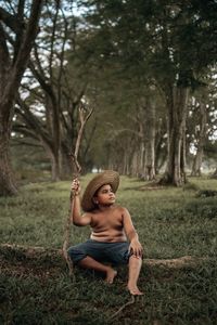 Shirtless boy holding branch while sitting on field against trees