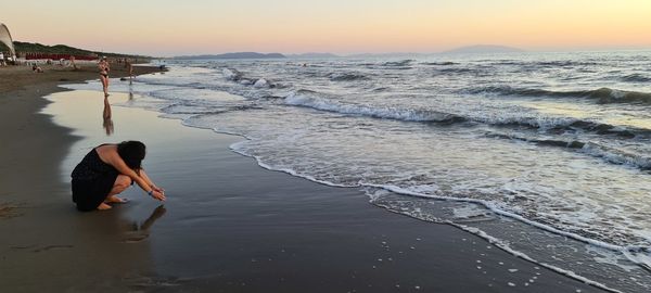 Rear view of woman standing at beach against sky during sunset