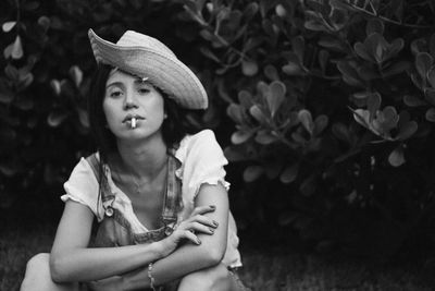 Portrait of young woman in hat smoking cigarette against plants
