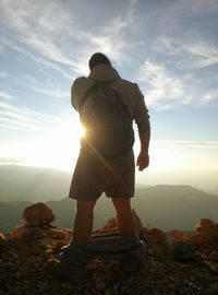 Rear view of hiker removing pants on mountain against sky during sunset