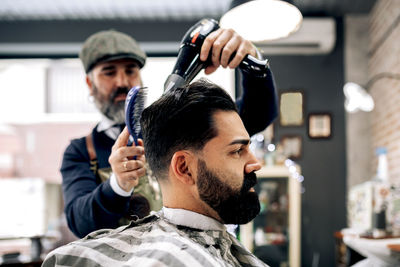 Concentrated mature ethnic barber styling dark hair of serious brutal client with dryer and comb in modern barbershop in daylight