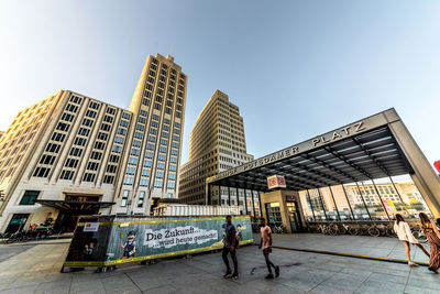 People walking by modern buildings against clear sky