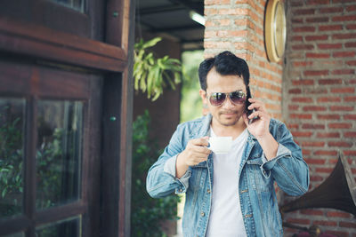 Young man holding coffee cup while using smart phone against brick wall at cafe