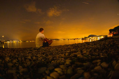 Side view of man sitting at lakeshore during sunset