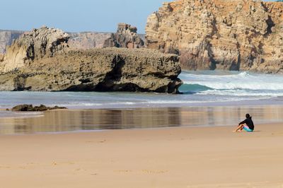 Woman sitting on sea shore by rock formation against sky