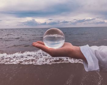 Cropped hand of young woman holding crystal ball over sea against cloudy sky during sunset