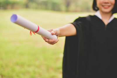 Midsection of young woman in graduation gown holding certificate while standing at park