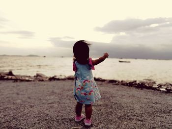 Rear view of baby girl standing at beach against sky during sunset
