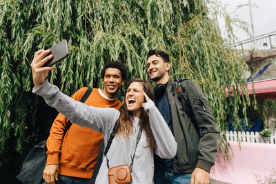 Smiling friends taking selfie with mobile phone while standing by tree in city