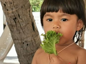 Close-up of shirtless baby girl eating vegetable against tree trunk