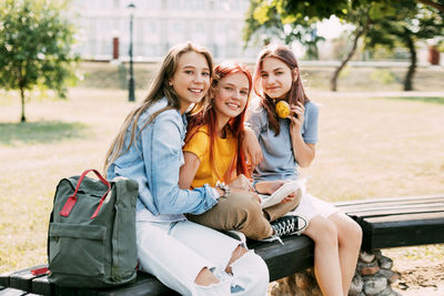 Three schoolgirls are sitting on a park bench, talking, preparing together with the lesson. 