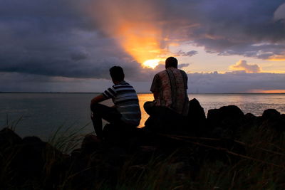 Men sitting on beach against sky during sunset