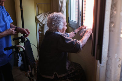 Side view of crop anonymous nurse in uniform standing near sick elderly female sitting in wheelchair near window in light room