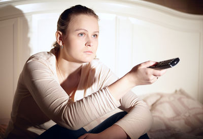 Woman holding remote control while sitting on bed at home