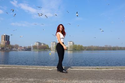 Full length portrait of young woman standing by lake against sky
