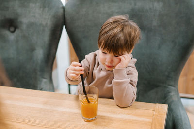 Four year old boy sits at a table in a cafe and looks at a glass of orange juice, holding a straw