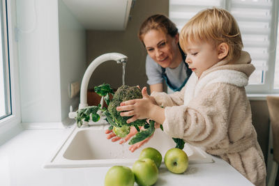 A mother and a small child are washing broccoli together in the kitchen for preparing dinner.