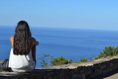 Rear view of woman with long black hair sitting against sea and clear blue sky