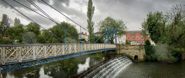 Bridge over river amidst trees and buildings against sky