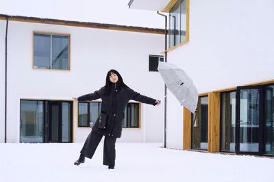 Portrait of mid adult woman holding umbrella while standing on snow covered field against building