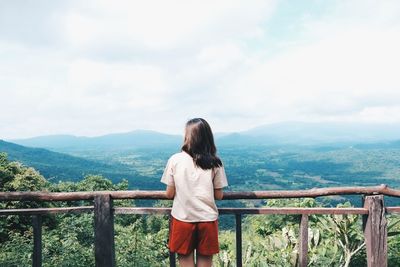Rear view of young woman looking at landscape while standing by railing against cloudy sky