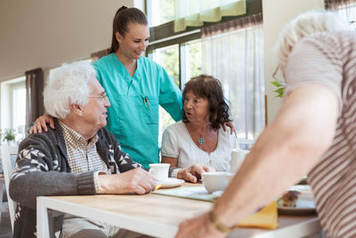 Smiling nurse listening to conversation of senior couple during breakfast at nursing home