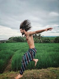 Side view of shirtless man jumping on grass against sky