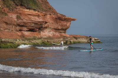 Man and woman paddleboarding in sea against rocky cliff