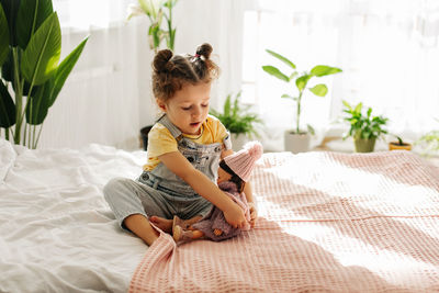 A little girl is sitting on the bed in the bedroom and playing with her doll, tying her scarf
